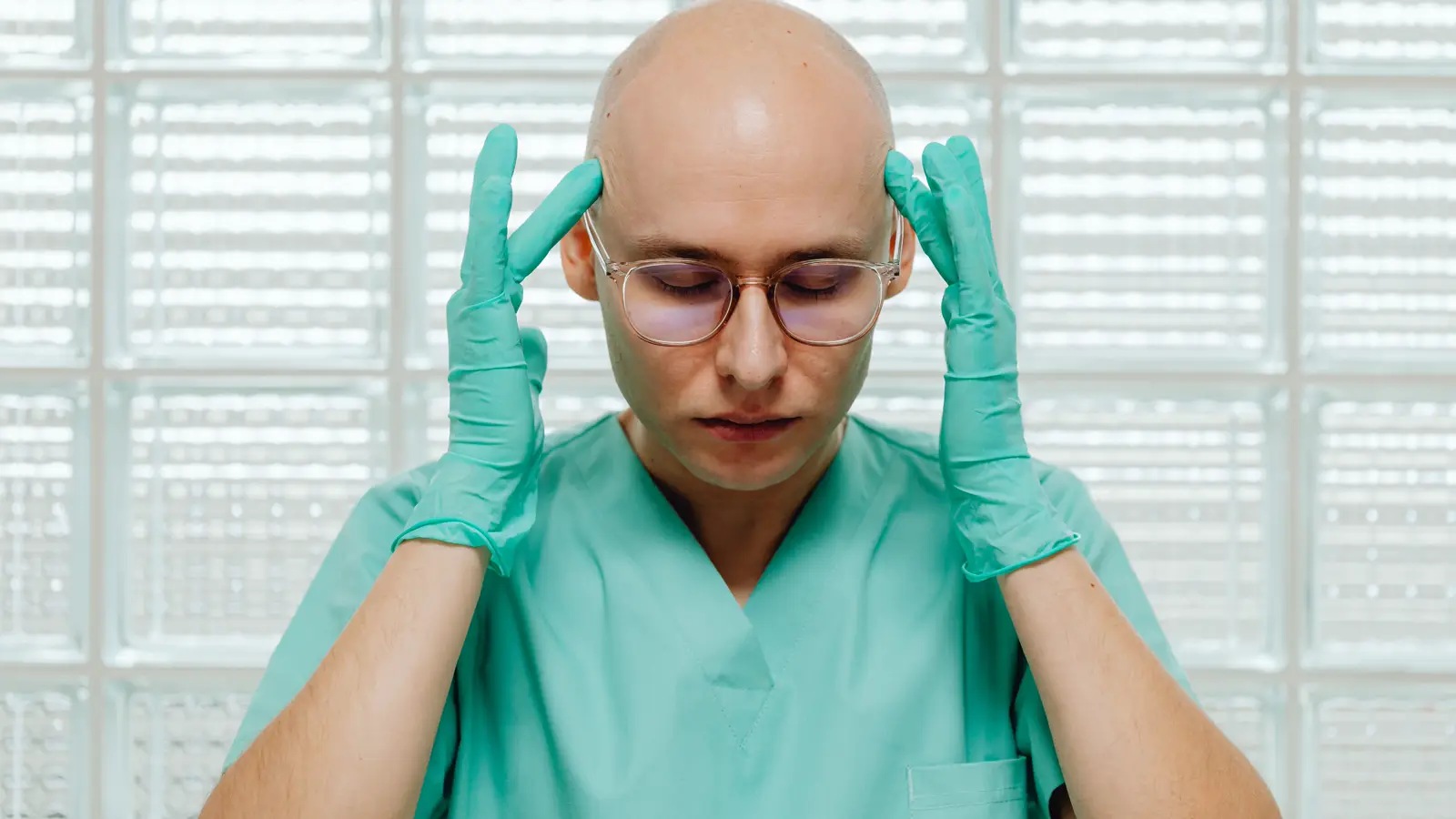Profissional de saúde com jaleco verde, óculos e luvas, de olhos fechados e cabeça baixa segurando as têmporas, retratando o desgaste de um caso de assédio moral no hospital