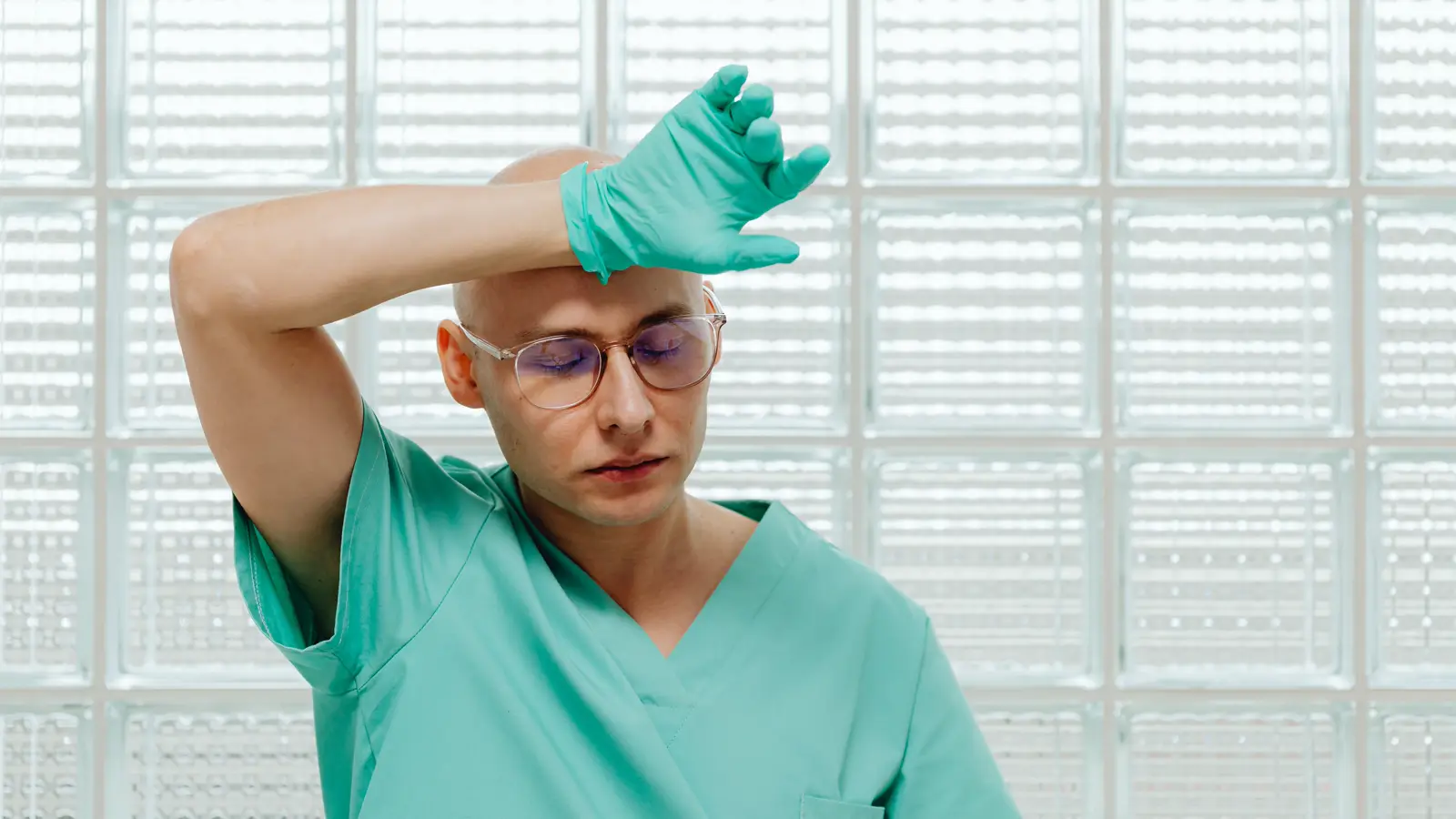Profissional de saúde de scrub verde com os olhos fechados e a testa apoiada na luva, exausto, em ambiente hospitalar, retratando o desgaste do assédio moral e sexual no ambiente hospitalar