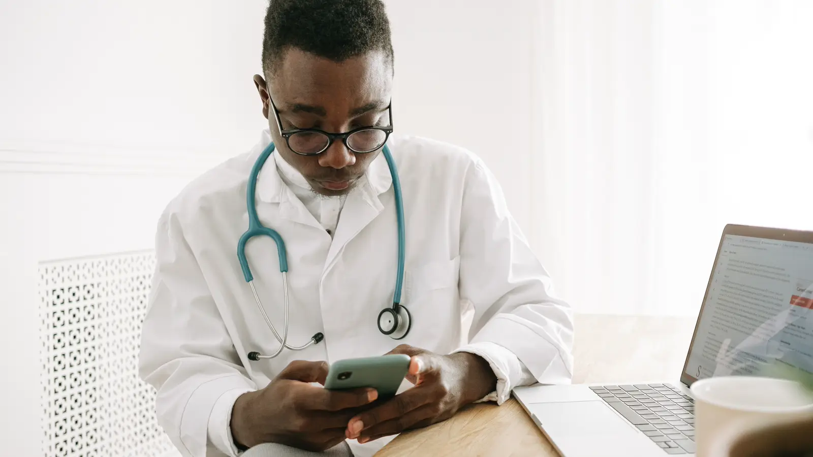 Médico de jaleco confere documento no celular ao lado do notebook, checando validade da assinatura eletrônica em receita e atestado digital