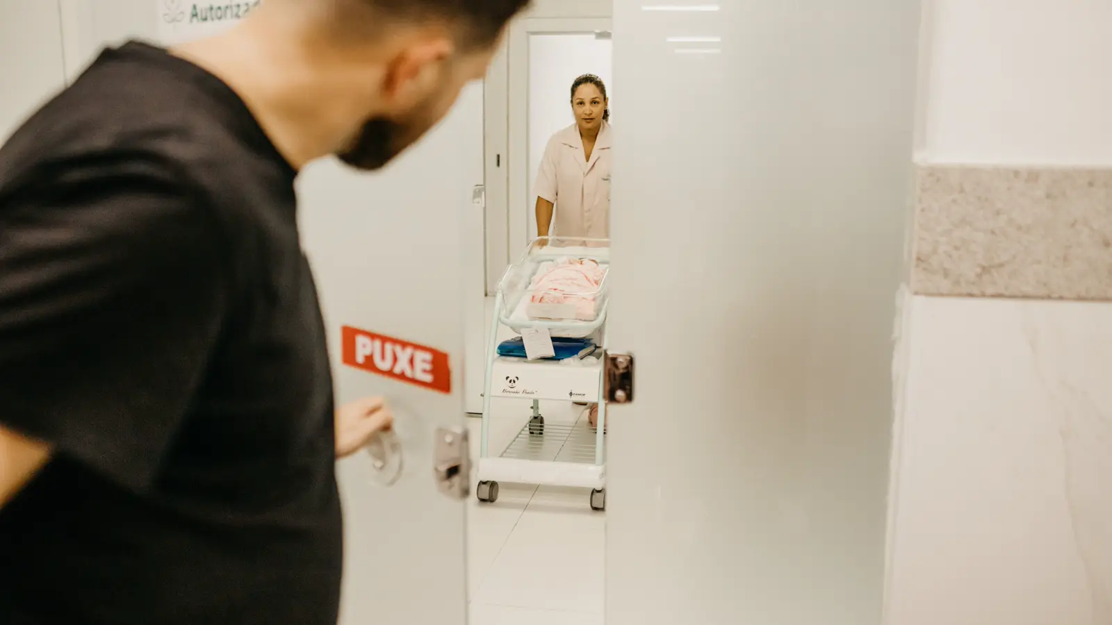 Pai de jaleco escuro parado em corredor de hospital enquanto enfermeira conduz berço com recém-nascido, ilustrando a licença-paternidade do médico