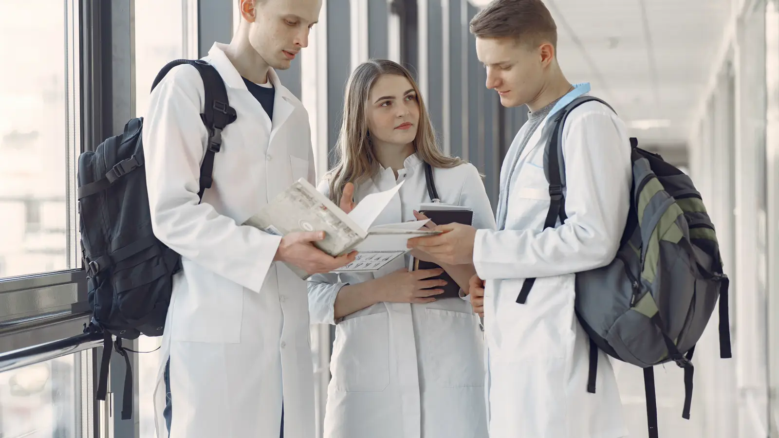 Três estudantes de medicina de jaleco branco conferindo anotações em corredor de faculdade, representando quem se formou em medicina fora do Brasil e vai prestar o Revalida