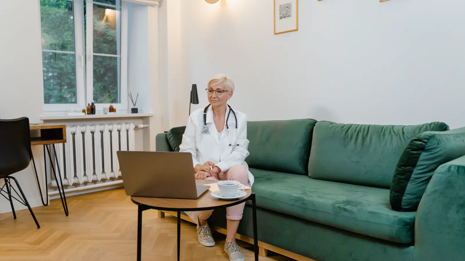 Médica de jaleco e estetoscópio faz teleconsulta pelo notebook, sentada em um sofá fora do consultório