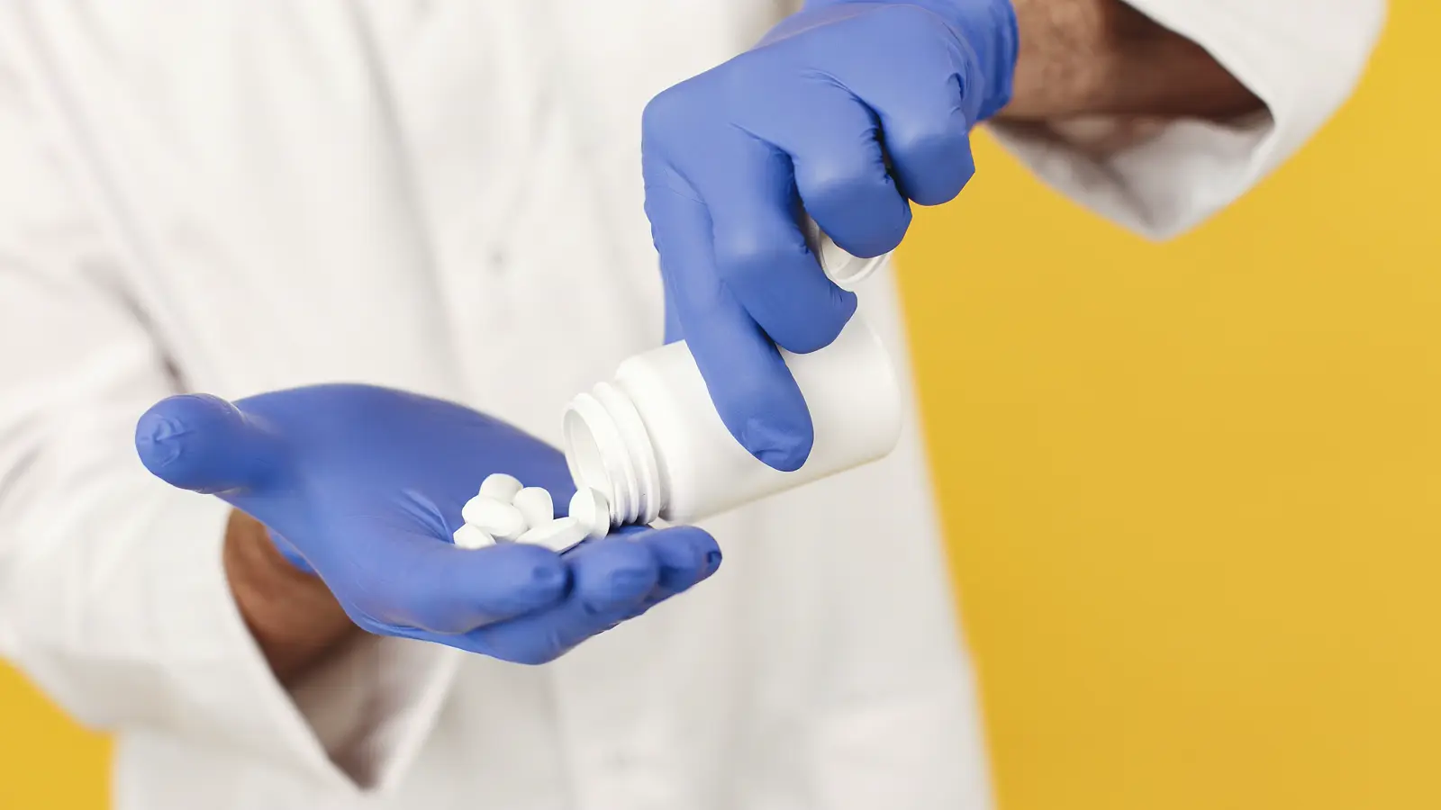 Profissional de jaleco e luvas despejando comprimidos de um frasco na palma da mão, representando a prescrição e dispensação de medicamento