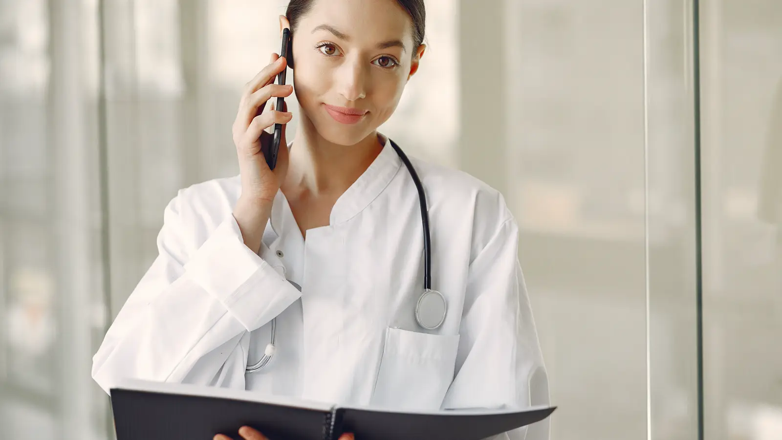 Médica de jaleco com estetoscópio falando ao telefone celular em corredor de hospital, ilustrando o sobreaviso por WhatsApp fora do plantão