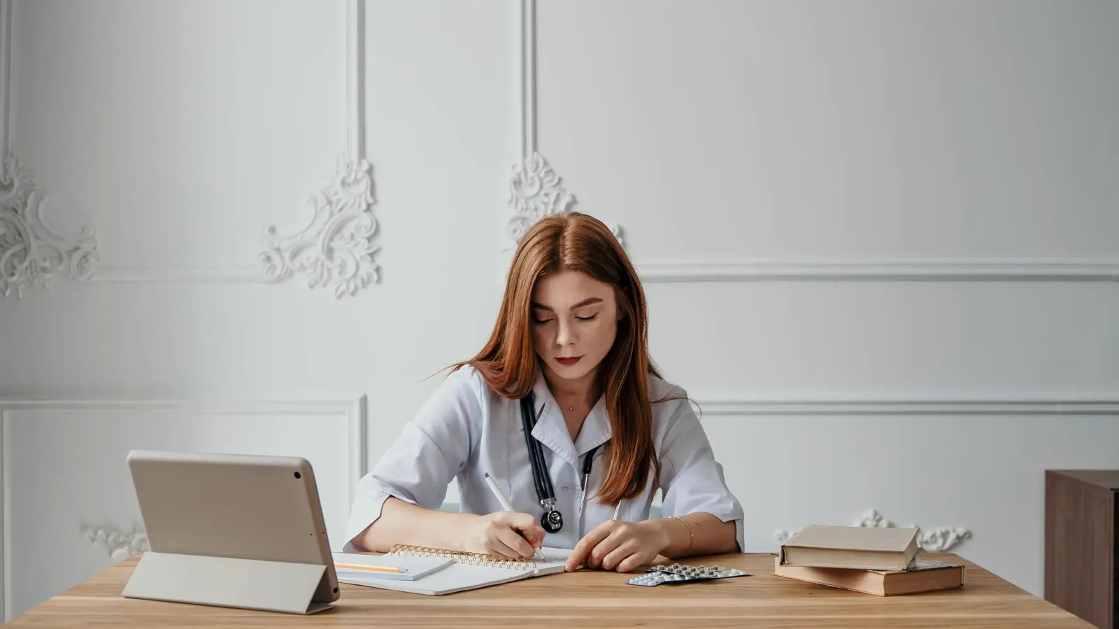 Médica de jaleco branco escrevendo em um caderno à mesa, com tablet e livros ao lado, ilustrando a decisão de mudar de especialidade