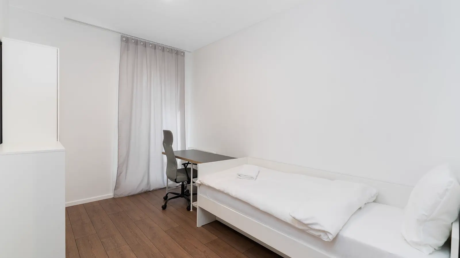 Quarto simples de alojamento com cama de solteiro e escrivaninha, moradia do médico residente