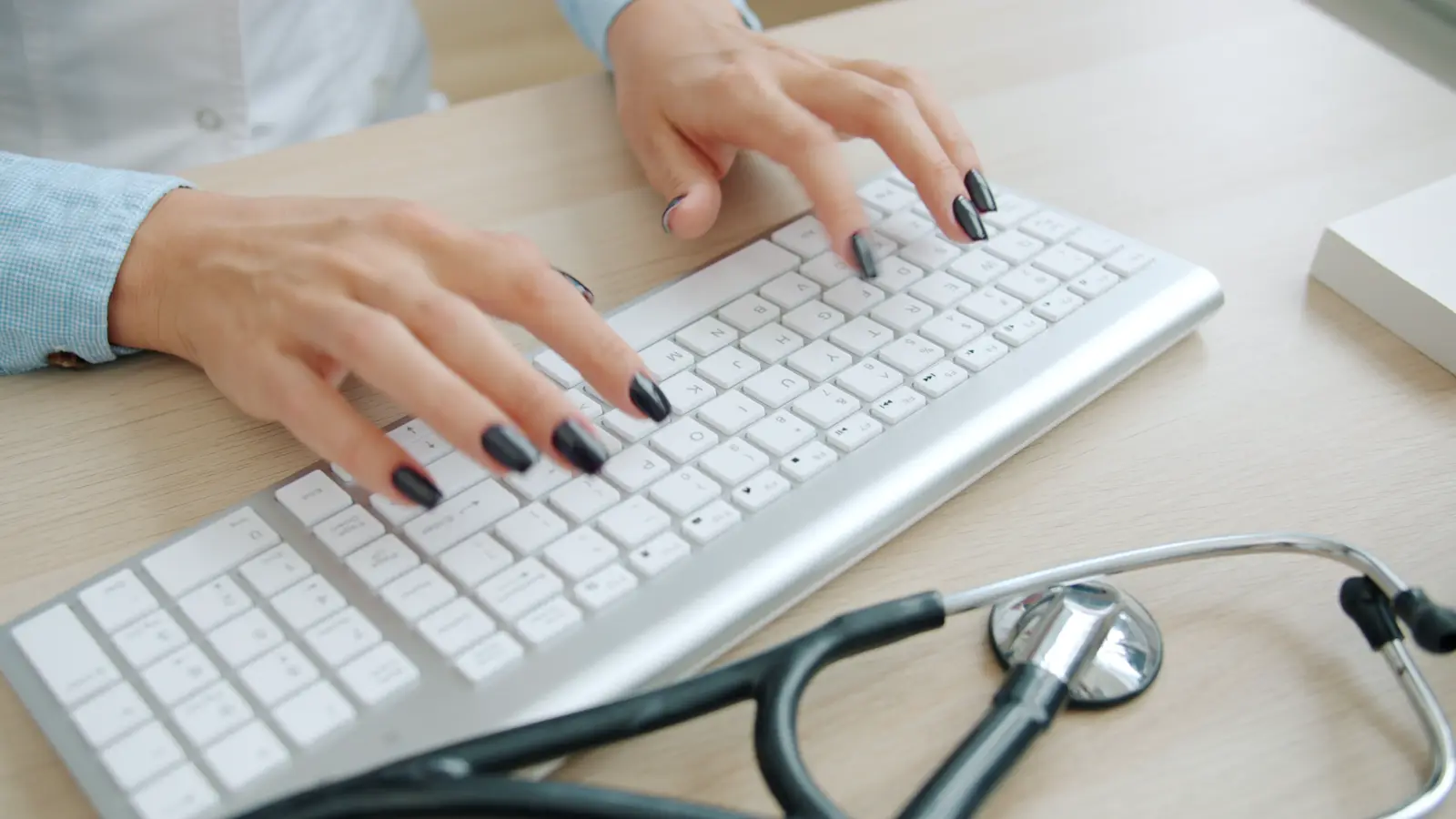 Mãos de profissional de saúde digitando em teclado ao lado de estetoscópio sobre mesa, ilustrando o uso de sistema de prontuário eletrônico no consultório